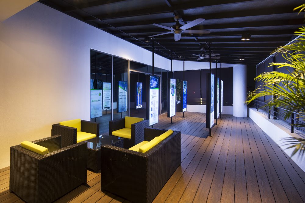 Backlit signage panels line a covered outdoor terrace, furnished with woven lounge seating and framed by tropical planting.