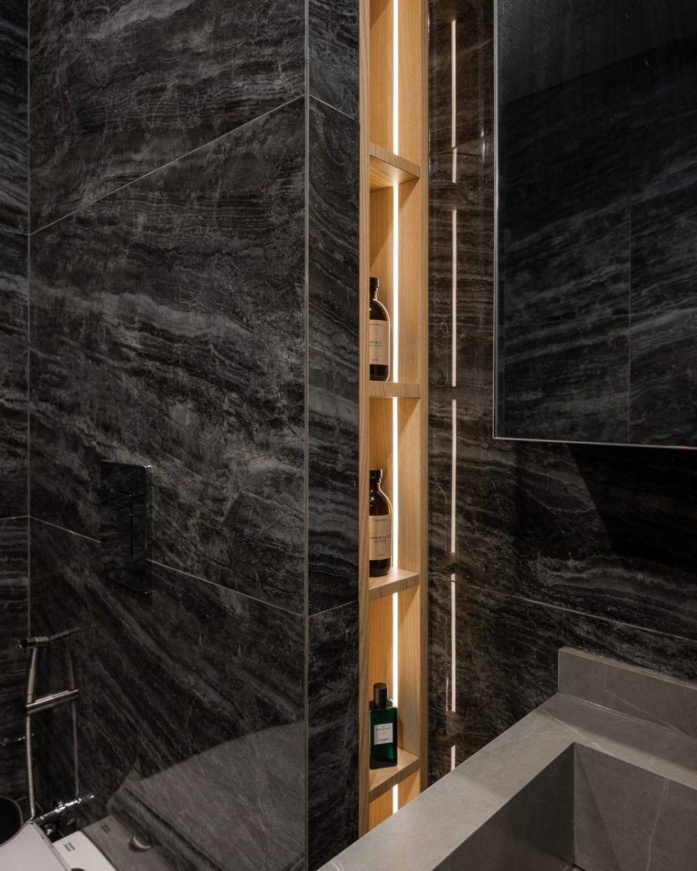 A vertical backlit niche holds toiletries within easy reach of the shower, built into the dark stone wall.