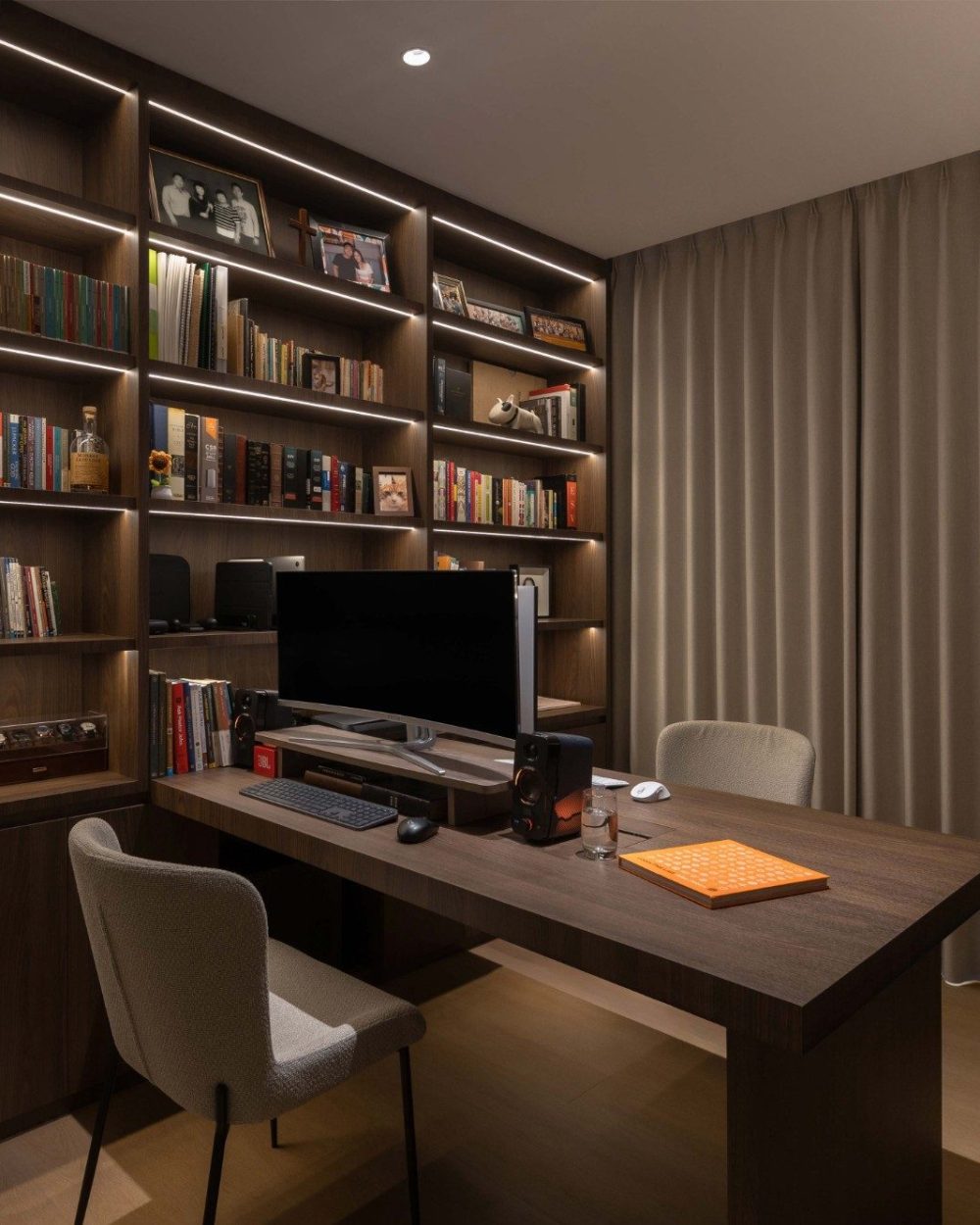 Full-height shelving is lit from within with a continuous LED strip, framing books, keepsakes and a watch winder box around a dual-monitor desk setup.