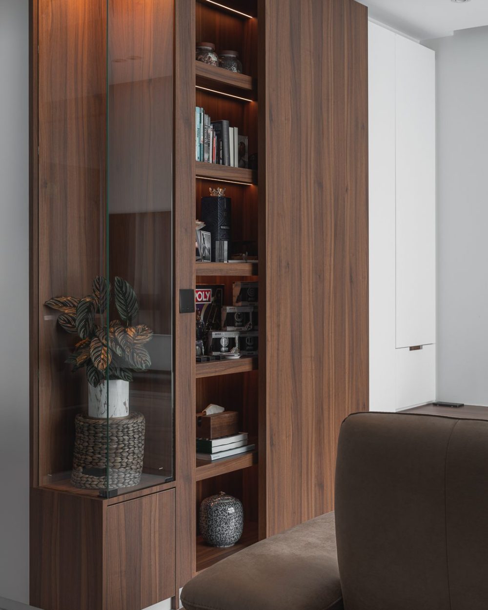 A tall glass-fronted cabinet holds books, whisky and personal keepsakes on backlit shelving, built into the living room's storage wall.