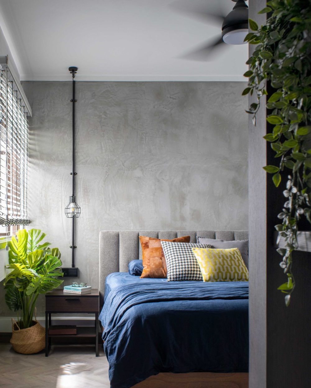 A large monstera plant and a woven basket soften the bedroom's industrial concrete wall, framed by a black ceiling fan.
