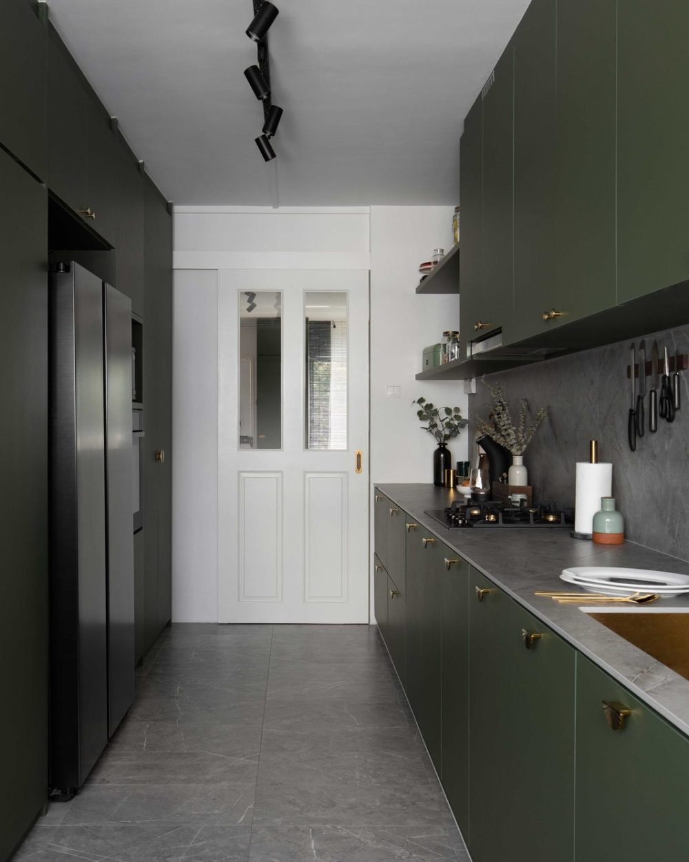 Deep olive green cabinetry with scalloped brass hardware wraps this galley kitchen, paired with a grey stone backsplash and a brass sink and tap.