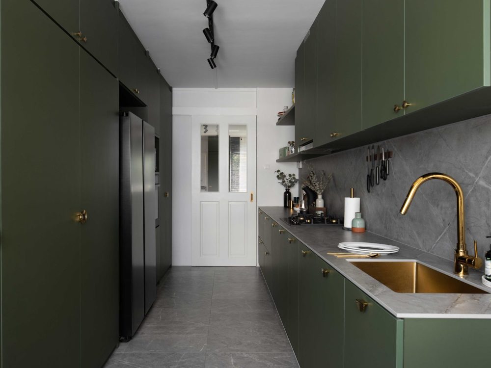 Deep olive green cabinetry with scalloped brass hardware wraps this galley kitchen, paired with a grey stone backsplash and a brass sink and tap.