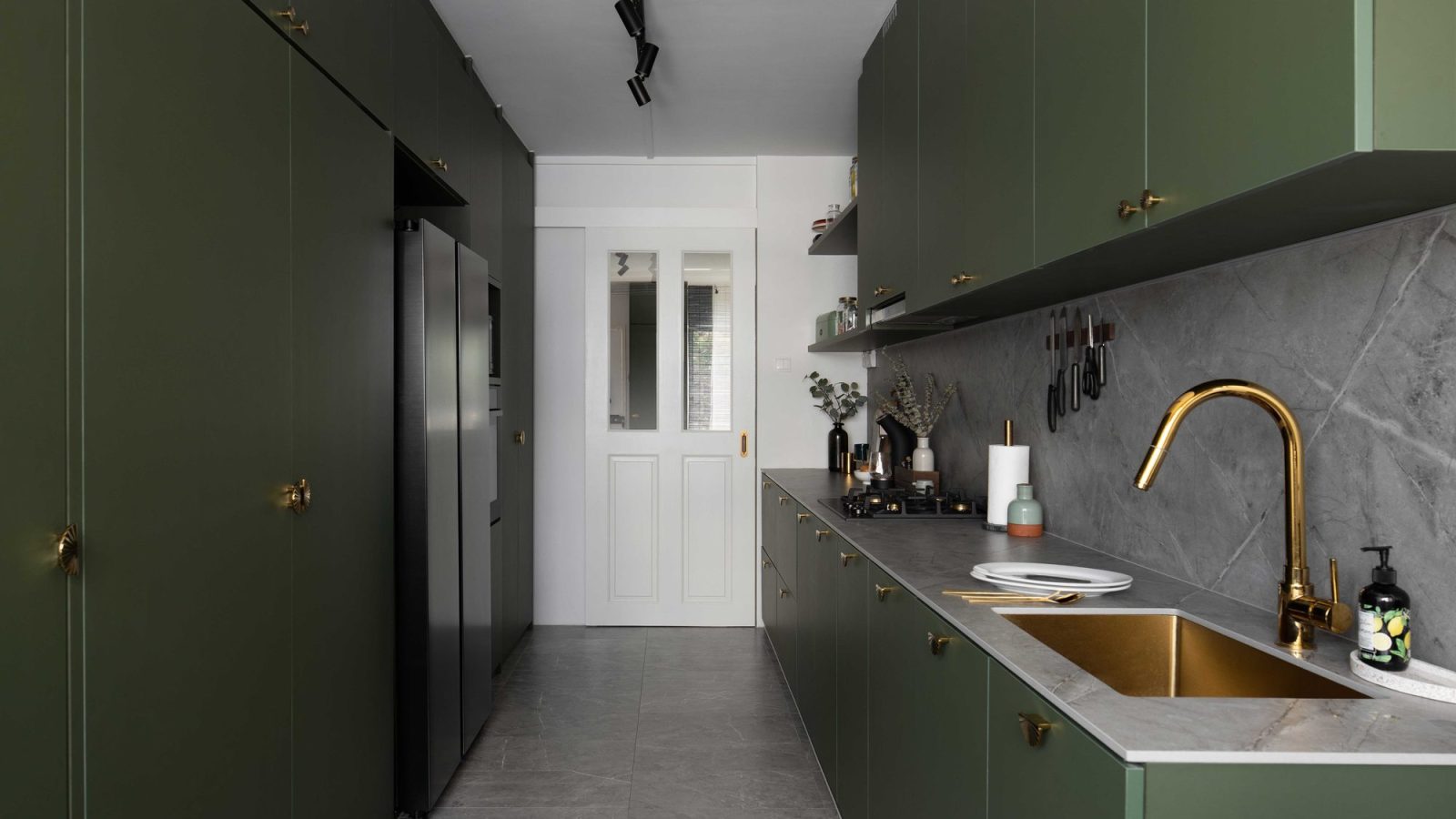 Deep olive green cabinetry with scalloped brass hardware wraps this galley kitchen, paired with a grey stone backsplash and a brass sink and tap.