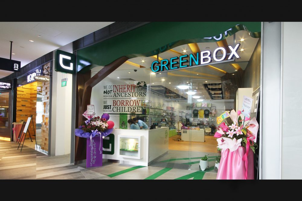 A curved green awning and backlit signage announce Greenbox's frontage, with a sculptural tree-branch motif framing the glass entrance.