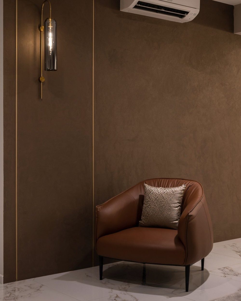 A brown leather armchair sits beneath a sculptural brass wall sconce, framed by a brass-inlaid textured wall panel and the air-conditioning unit above