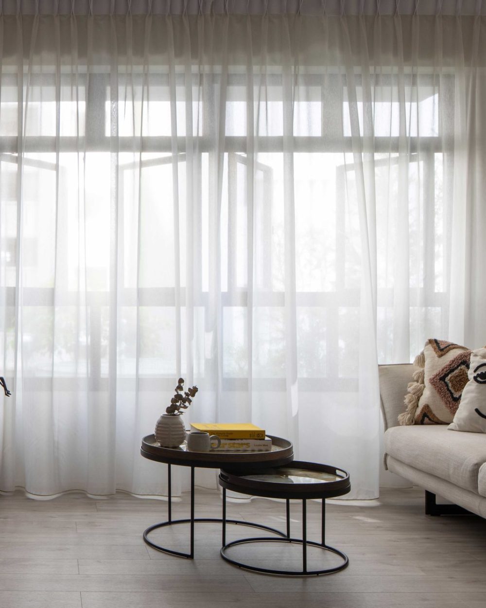 Sheer curtains soften the light across a plush cream sofa styled with playful embroidered cushions, framed by a warm timber sideboard.