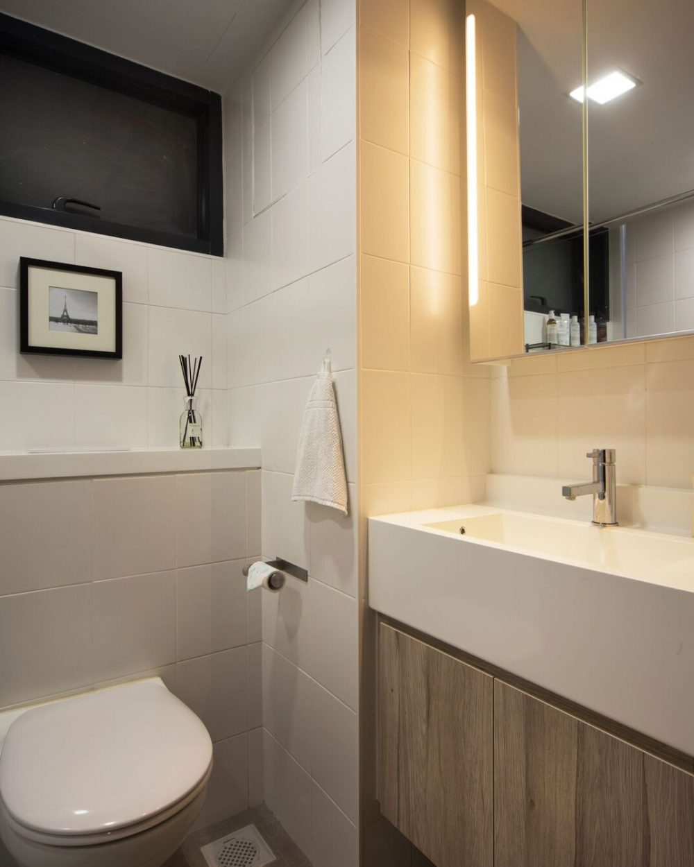 White wall tiles and a wood-toned vanity are lit by a vertical strip light built into the mirror cabinet, alongside a wall-hung toilet.