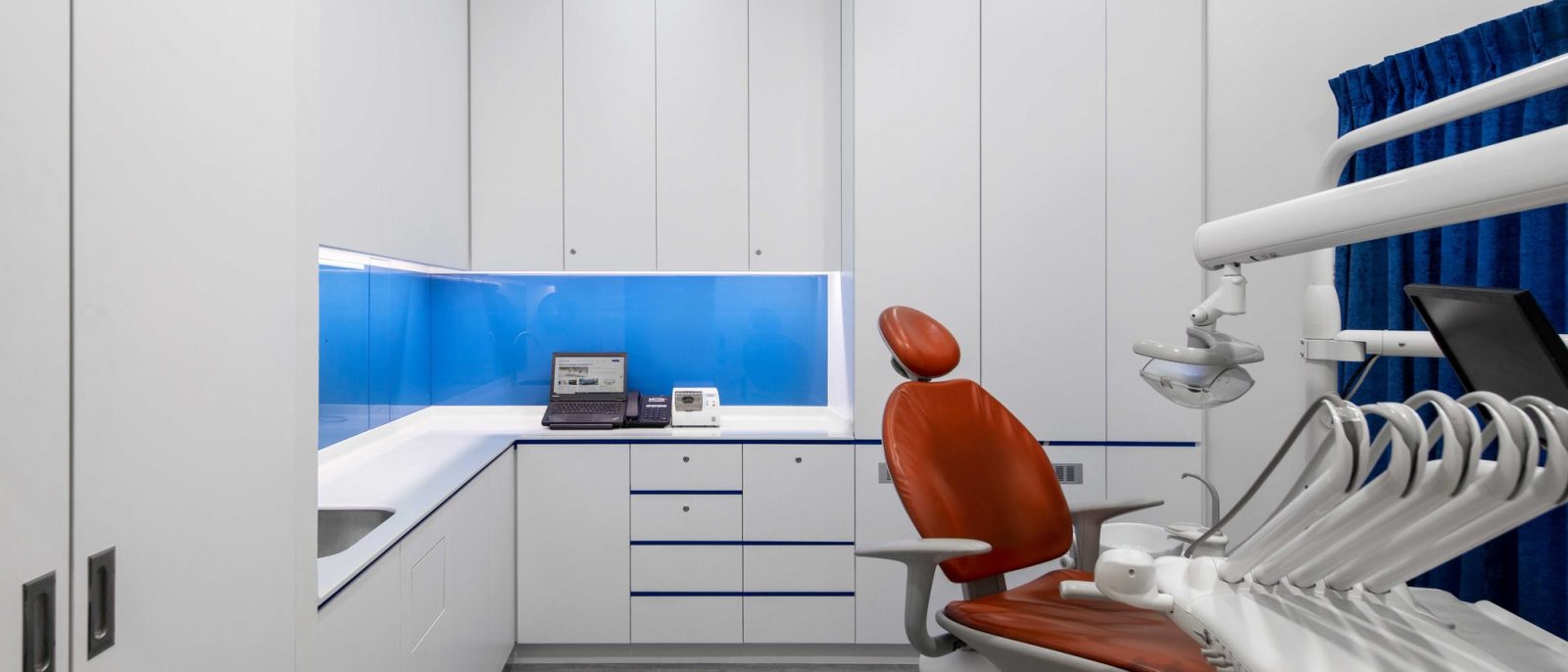A vivid blue glass backsplash and orange upholstered dental chair give this treatment room a bold, clinical-yet-approachable character.