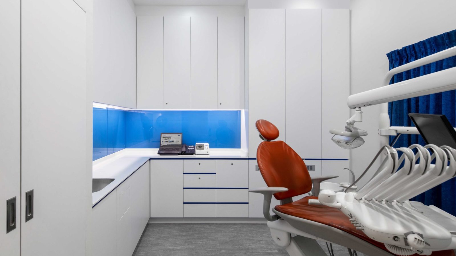 A vivid blue glass backsplash and orange upholstered dental chair give this treatment room a bold, clinical-yet-approachable character.
