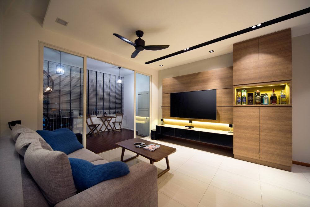 A backlit bar cabinet sits beside the TV wall, stocked with spirits and glassware, with the balcony's pendant lighting visible through the sliding doo