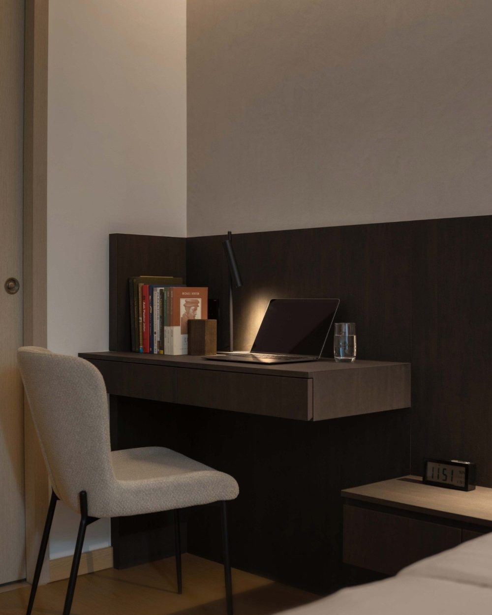 A slim built-in desk sits within the bedroom's timber-panelled wall, styled with a stack of books, a task lamp and a laptop — a quiet corner for work