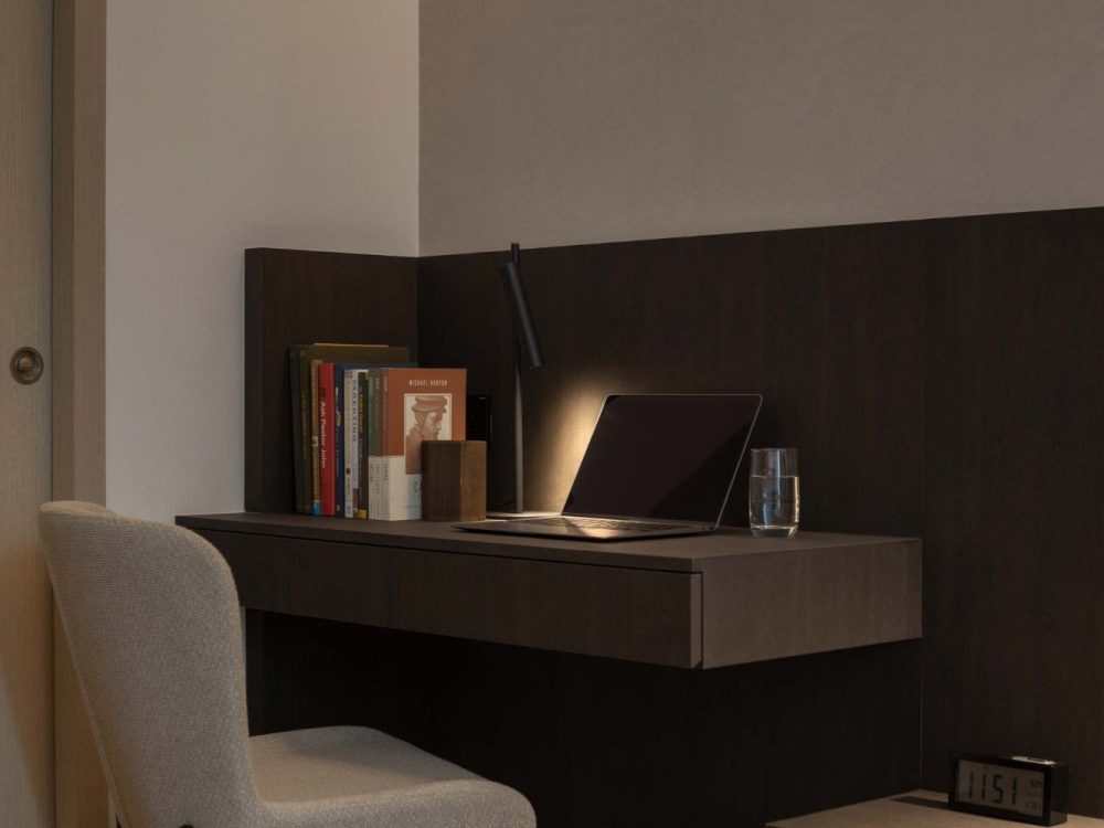 A slim built-in desk sits within the bedroom's timber-panelled wall, styled with a stack of books, a task lamp and a laptop — a quiet corner for work