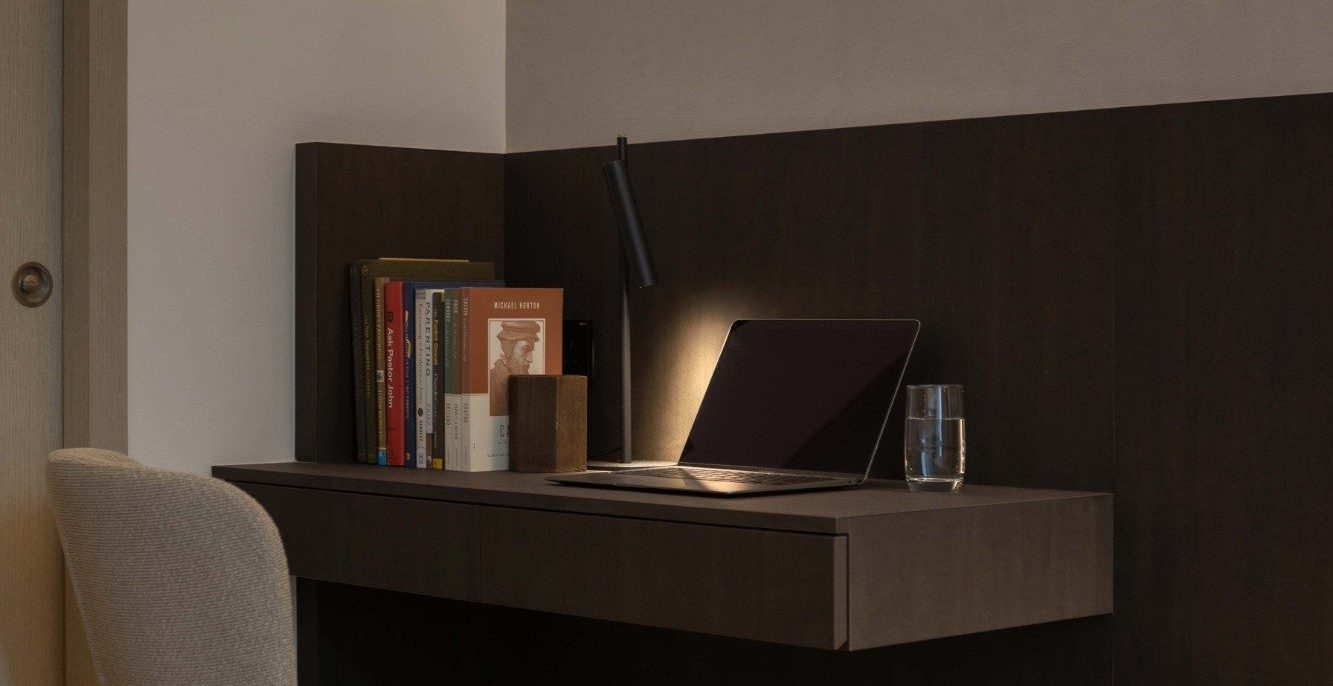 A slim built-in desk sits within the bedroom's timber-panelled wall, styled with a stack of books, a task lamp and a laptop — a quiet corner for work