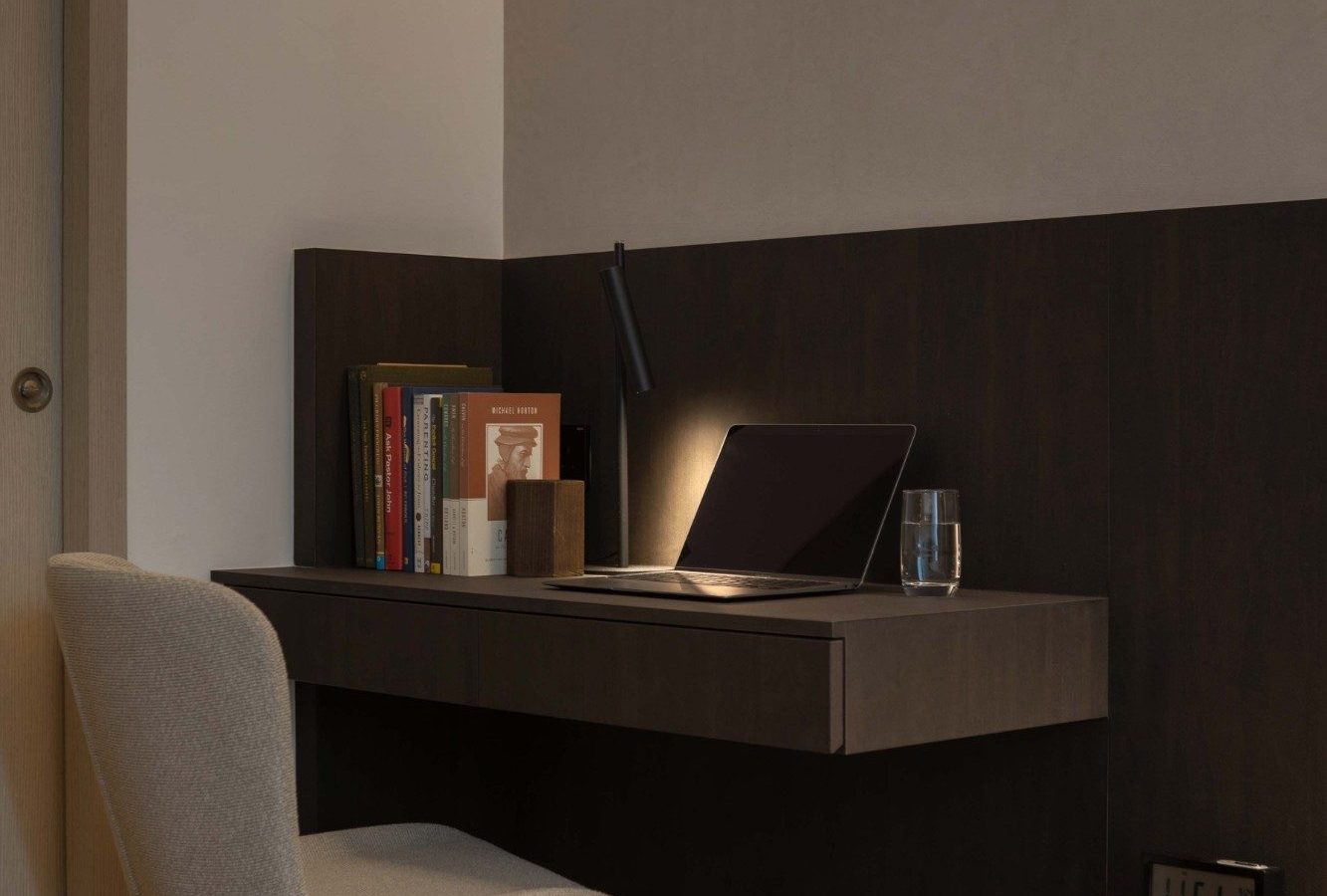 A slim built-in desk sits within the bedroom's timber-panelled wall, styled with a stack of books, a task lamp and a laptop — a quiet corner for work