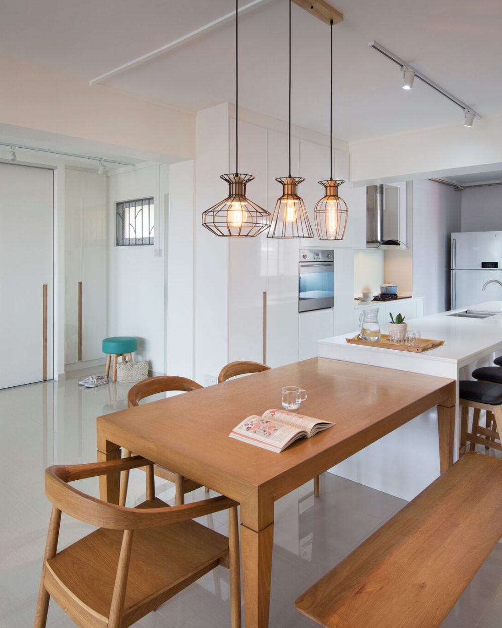 Three wire cage pendants hang over a bench-seated dining table, with a glossy white kitchen island and black bar stools completing the open-plan space
