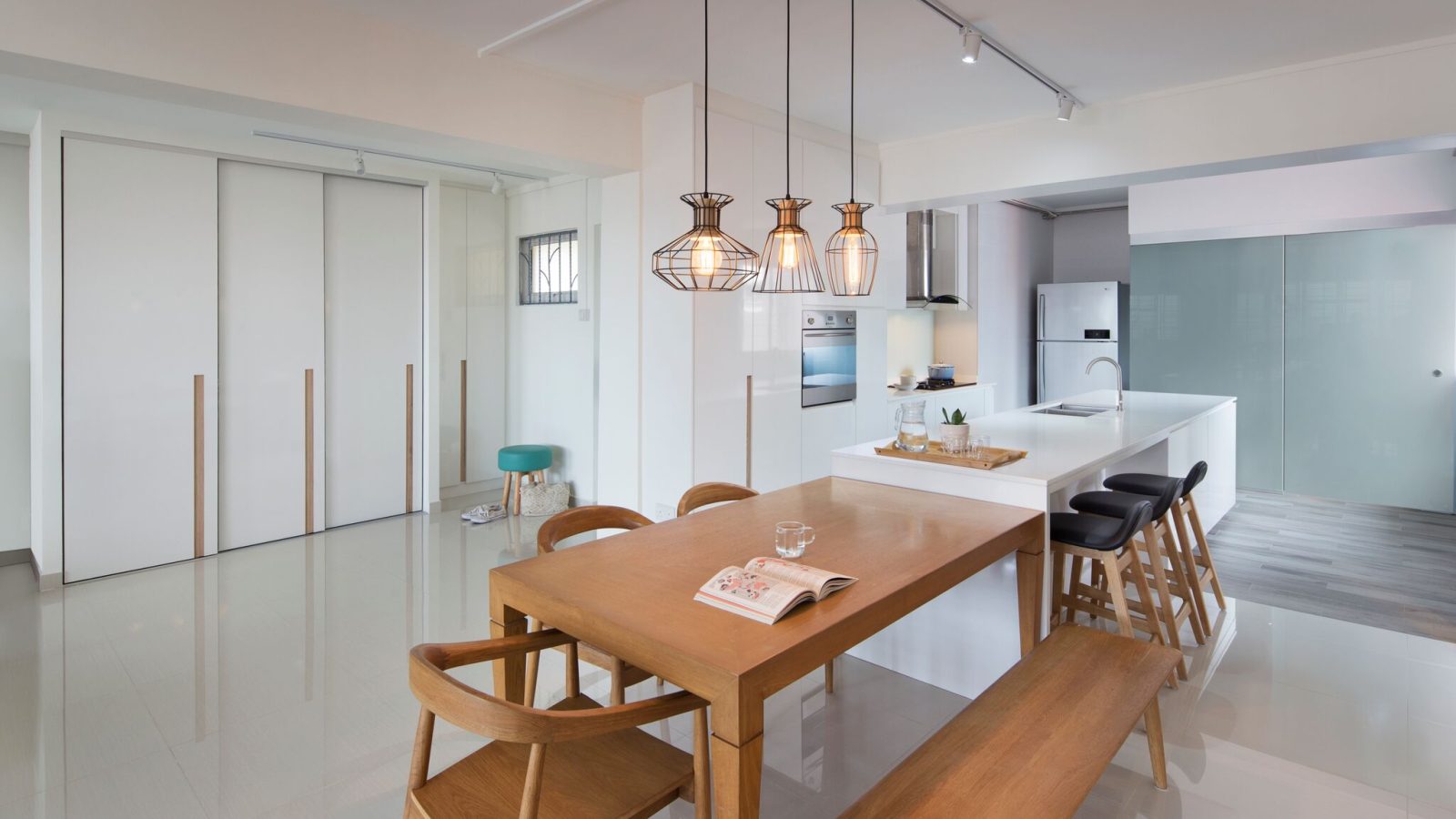 Three wire cage pendants hang over a bench-seated dining table, with a glossy white kitchen island and black bar stools completing the open-plan space