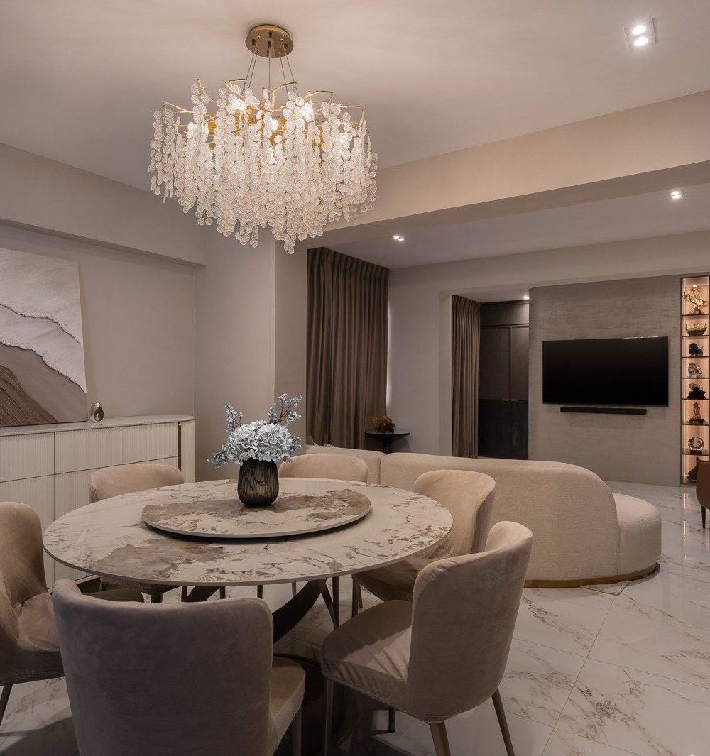 A crystal chandelier hangs over a round marble dining table, with a curved boucle sofa and a lit display-niche TV wall visible in the open-concept liv