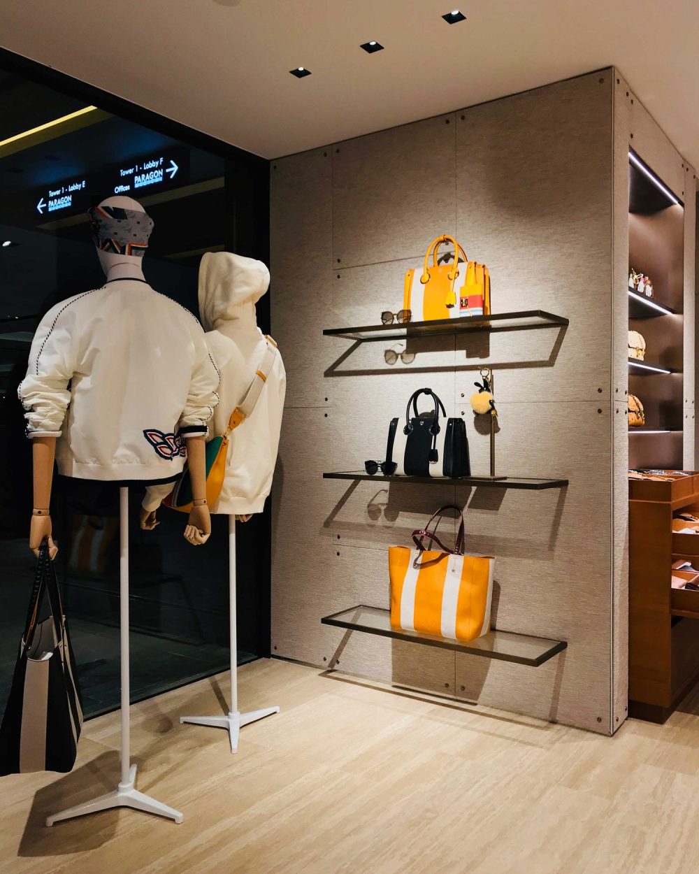 Two mannequins in cream outerwear beside a wall display of striped totes and bags.