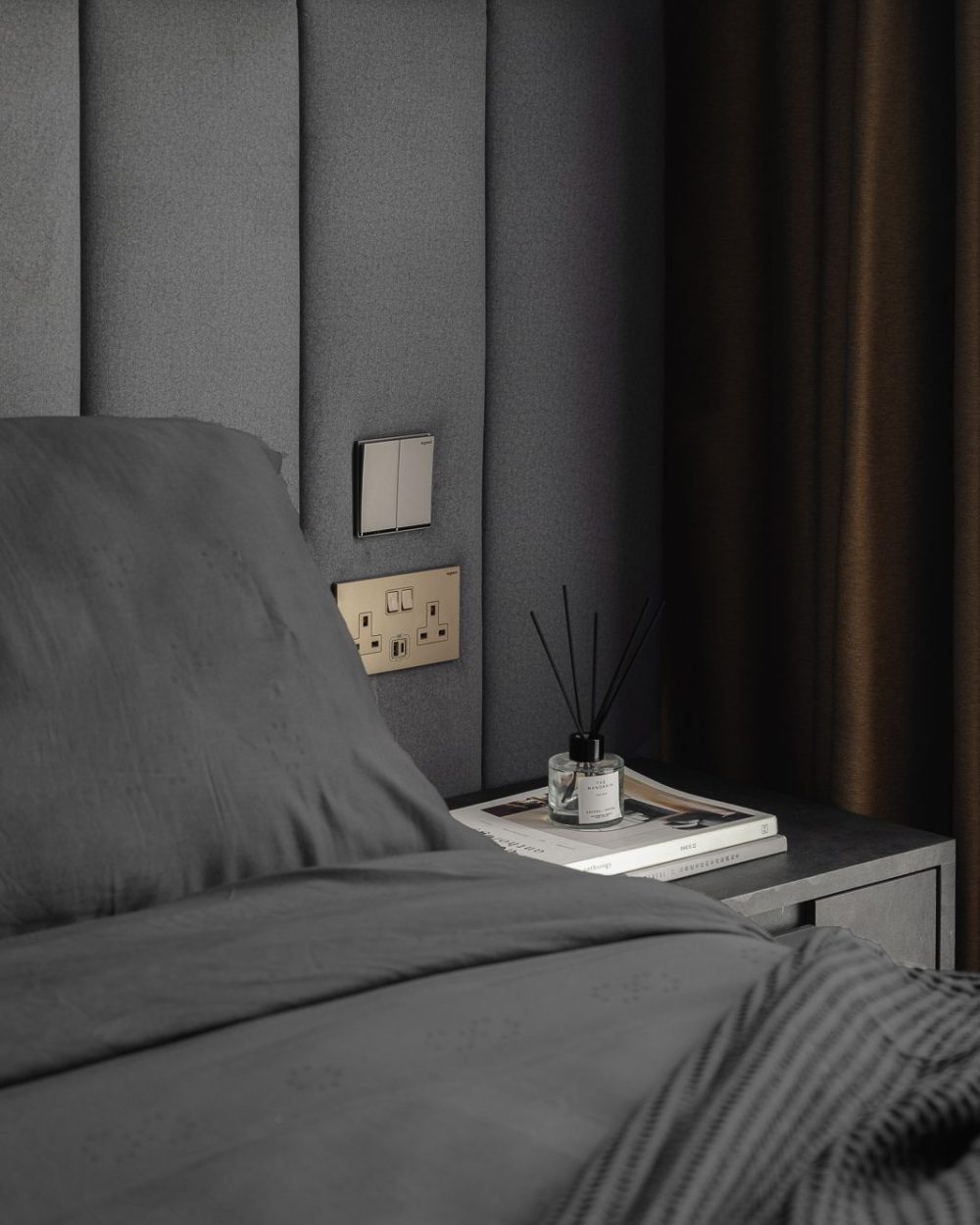 A reed diffuser and reading material sit on the bedside table, framed by brass-trimmed switches set into the upholstered wall.
