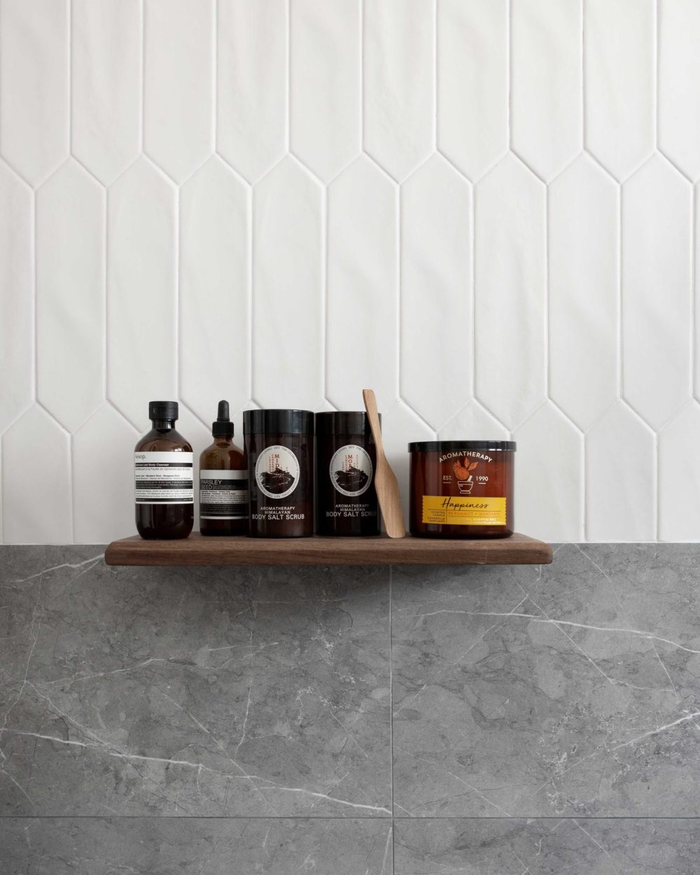 Skincare and bath products are displayed on a slim timber shelf, set against hexagonal white tile and grey marble.