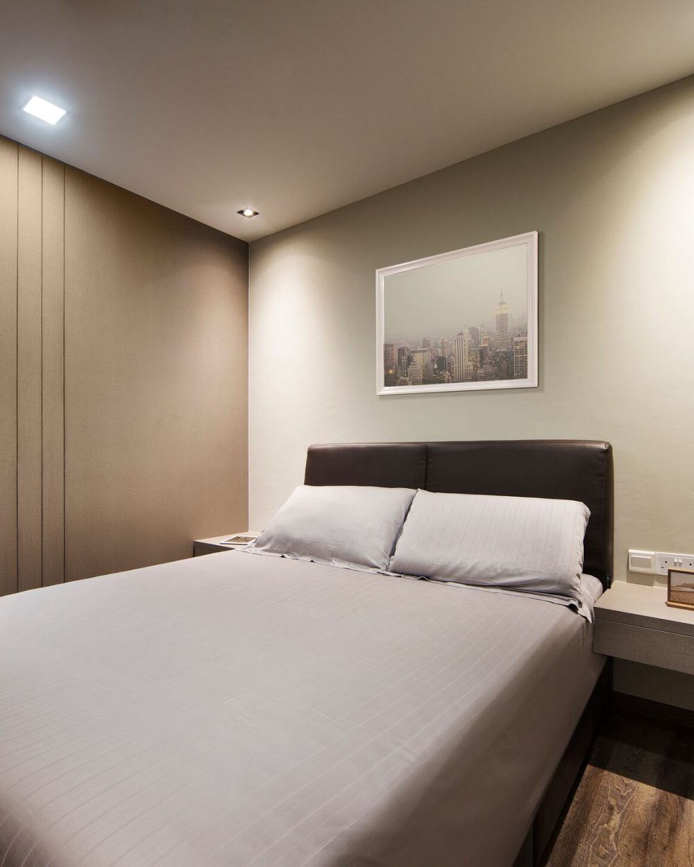 A brown leather headboard and floating bedside console sit beneath a framed cityscape print, against a soft sage-toned wall.