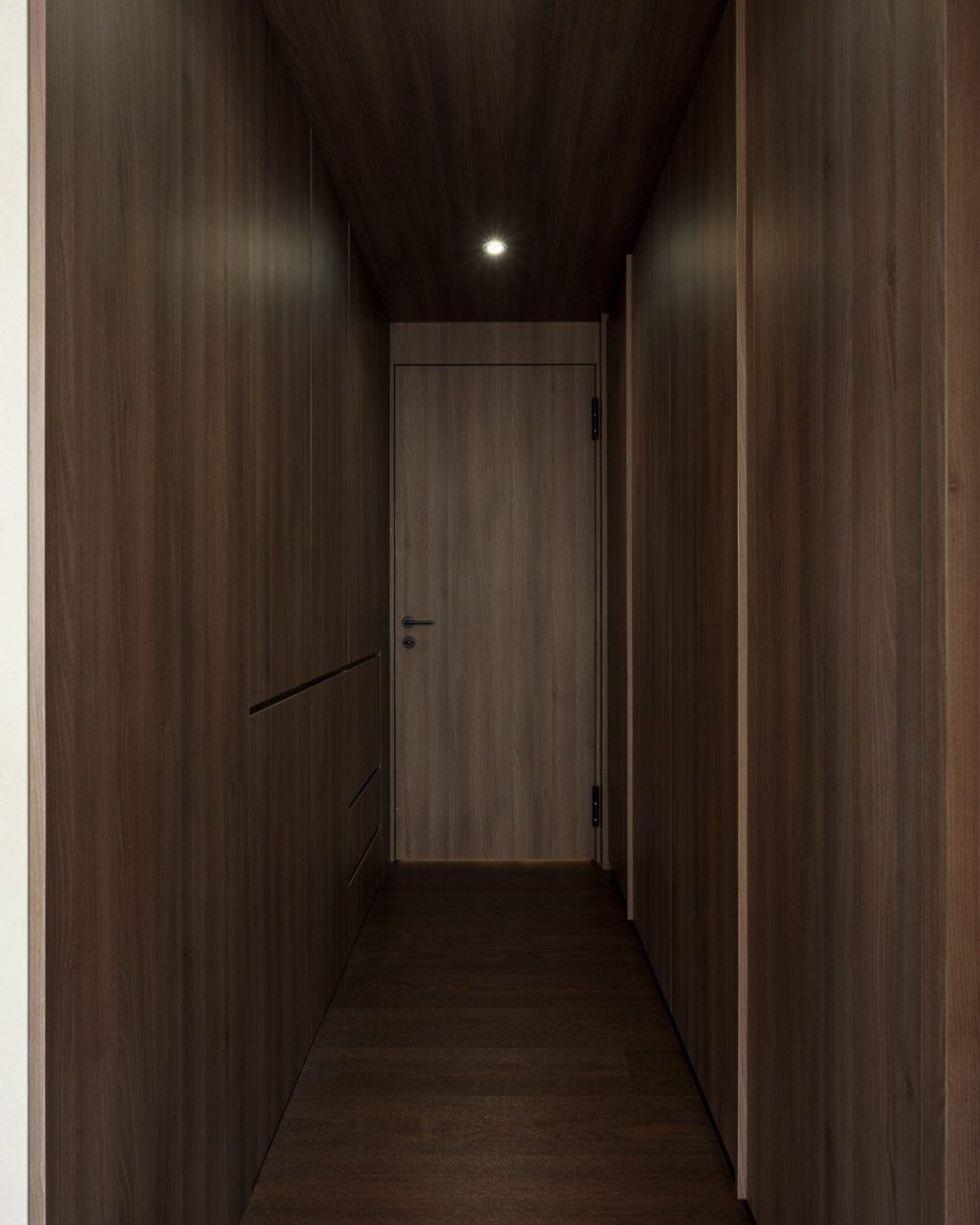 Floor-to-ceiling timber panelling and handle-less drawer fronts line this corridor, leading the eye toward a flush timber door lit by a single recesse