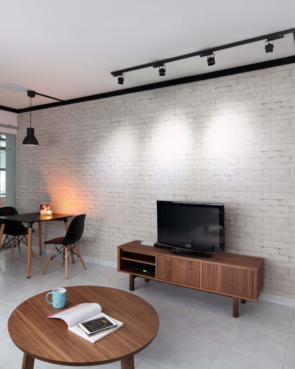 A raw-textured brick-effect wall anchors the living and dining space, framed by black track lighting and a slim walnut TV console. A glass-panelled sl
