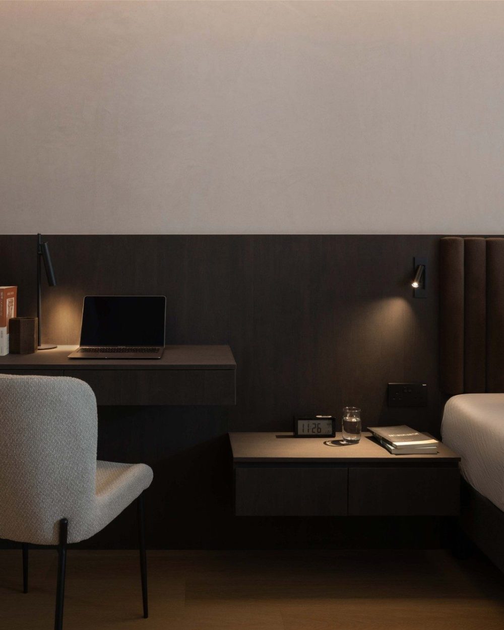 A channel-tufted velvet headboard anchors the bed against a slatted timber wall, with a wall-mounted reading light and a compact side table keeping th