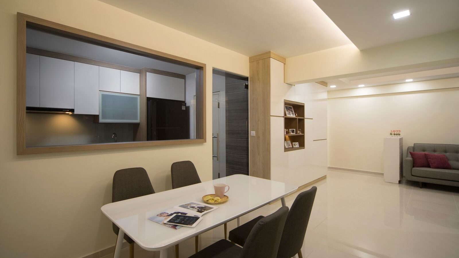 A timber-framed window connects the kitchen to the dining area, paired with a slim wood-and-white display shelf and a four-seat dining table.