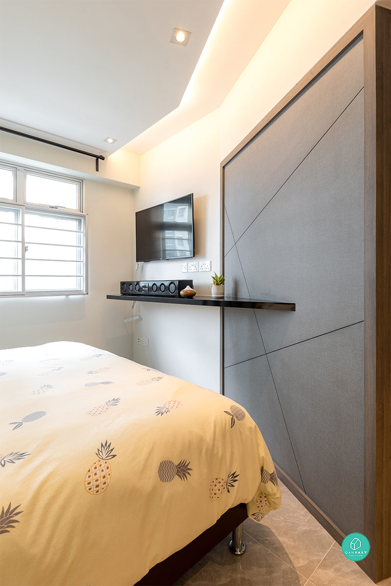 A grey upholstered headboard with a diamond stitch pattern anchors the bedroom, framed in warm timber and paired with a floating shelf.