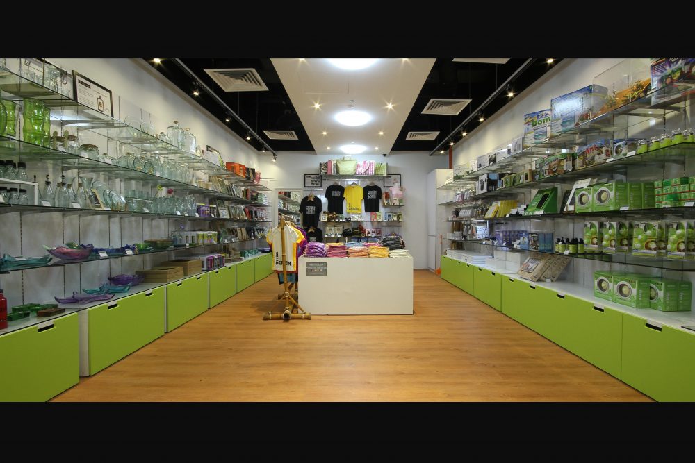Twin aisles of glass shelving and lime-green storage cabinetry display homeware and eco products along warm timber flooring.