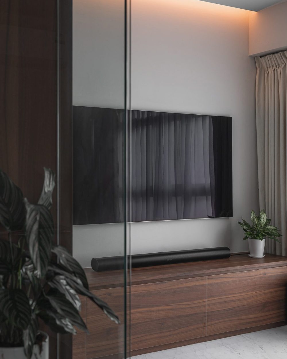 Seen through a glass pivot door, the TV wall's soft cove lighting and walnut media console glow warmly in the evening light.