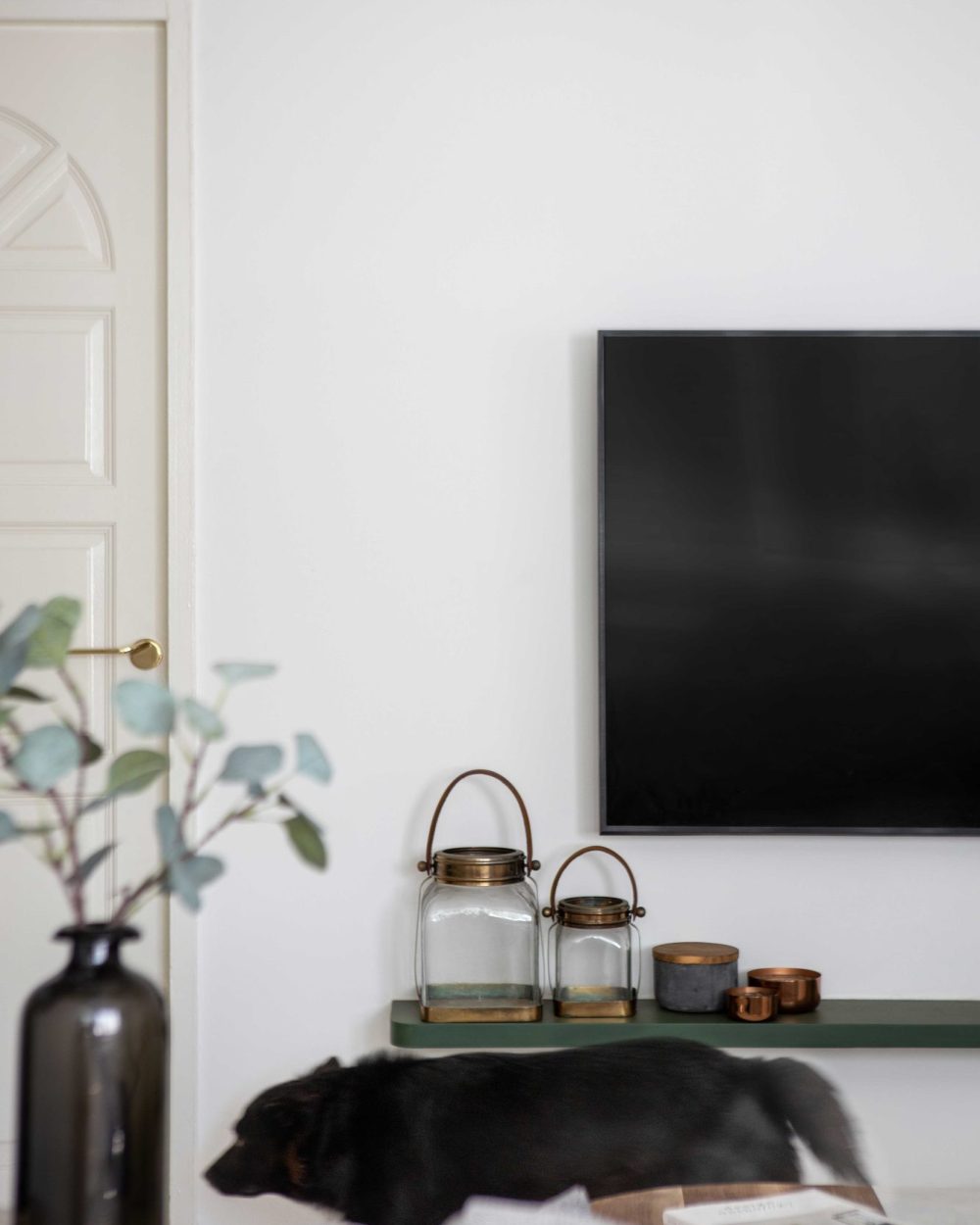 A slim green floating shelf holds brass lanterns and candles beneath the TV, styled simply and left uncluttered.