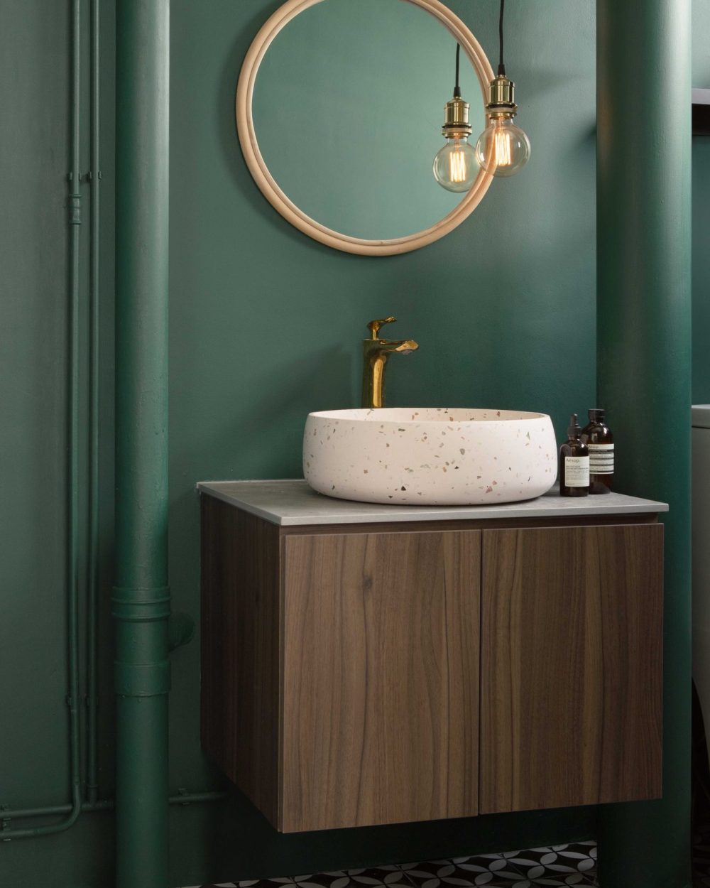 Deep green walls and painted exposed pipework frame a terrazzo vessel sink and brass tap in this striking ensuite bathroom.