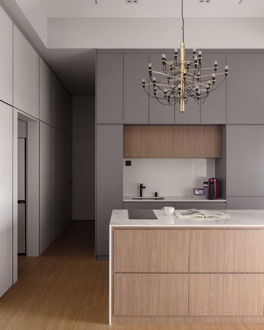 Matte grey cabinetry is broken up by a walnut-toned upper cabinet insert, with the same brass chandelier extending the dining area's lighting into the