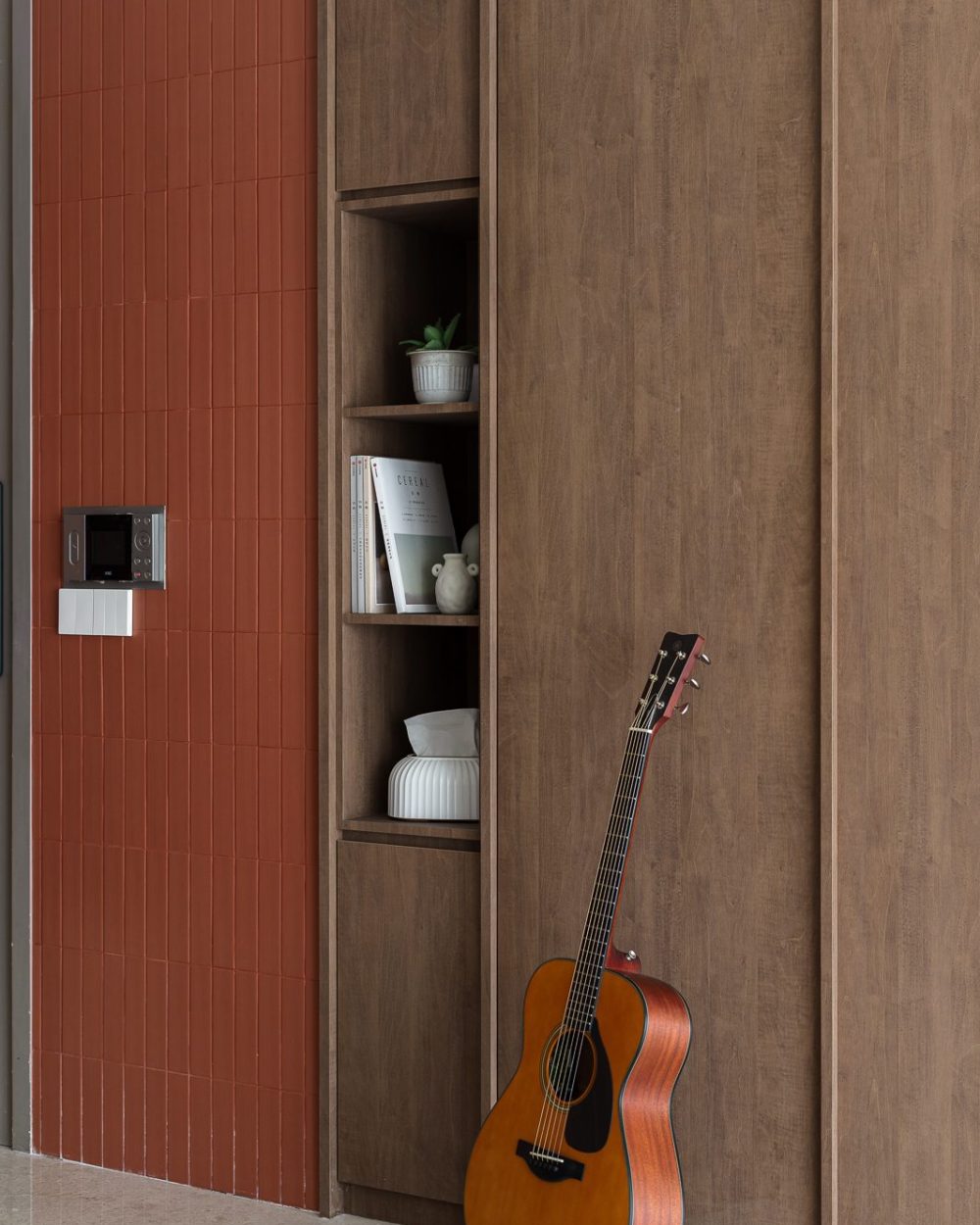 Open timber shelving displays books and ceramics beside a spot for an acoustic guitar, framed by the terracotta tile wall.