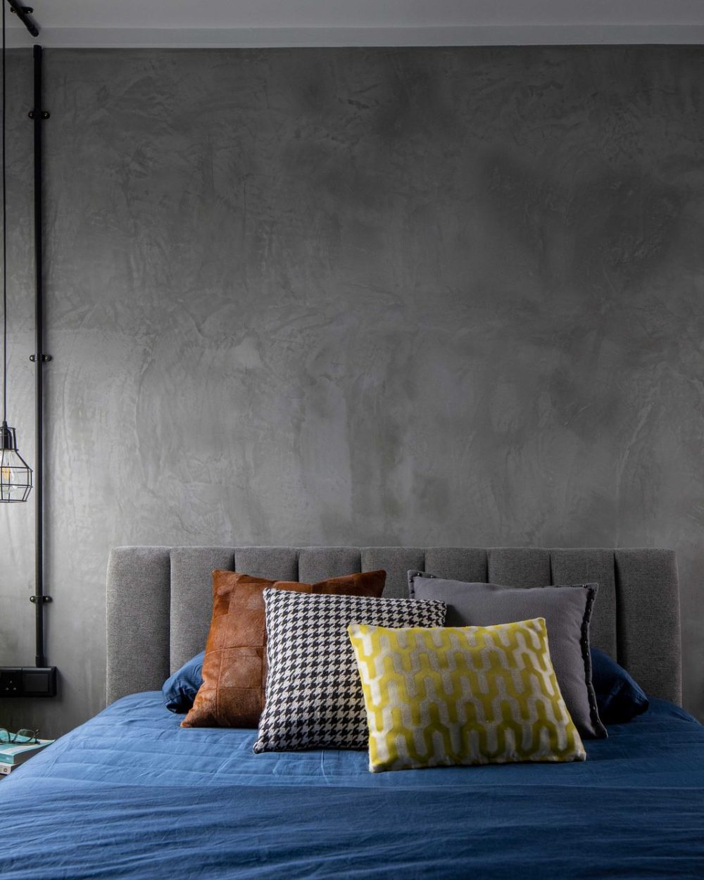 Raw concrete plaster wraps the headboard wall, flanked by exposed black conduit pipes ending in cage pendant lights.