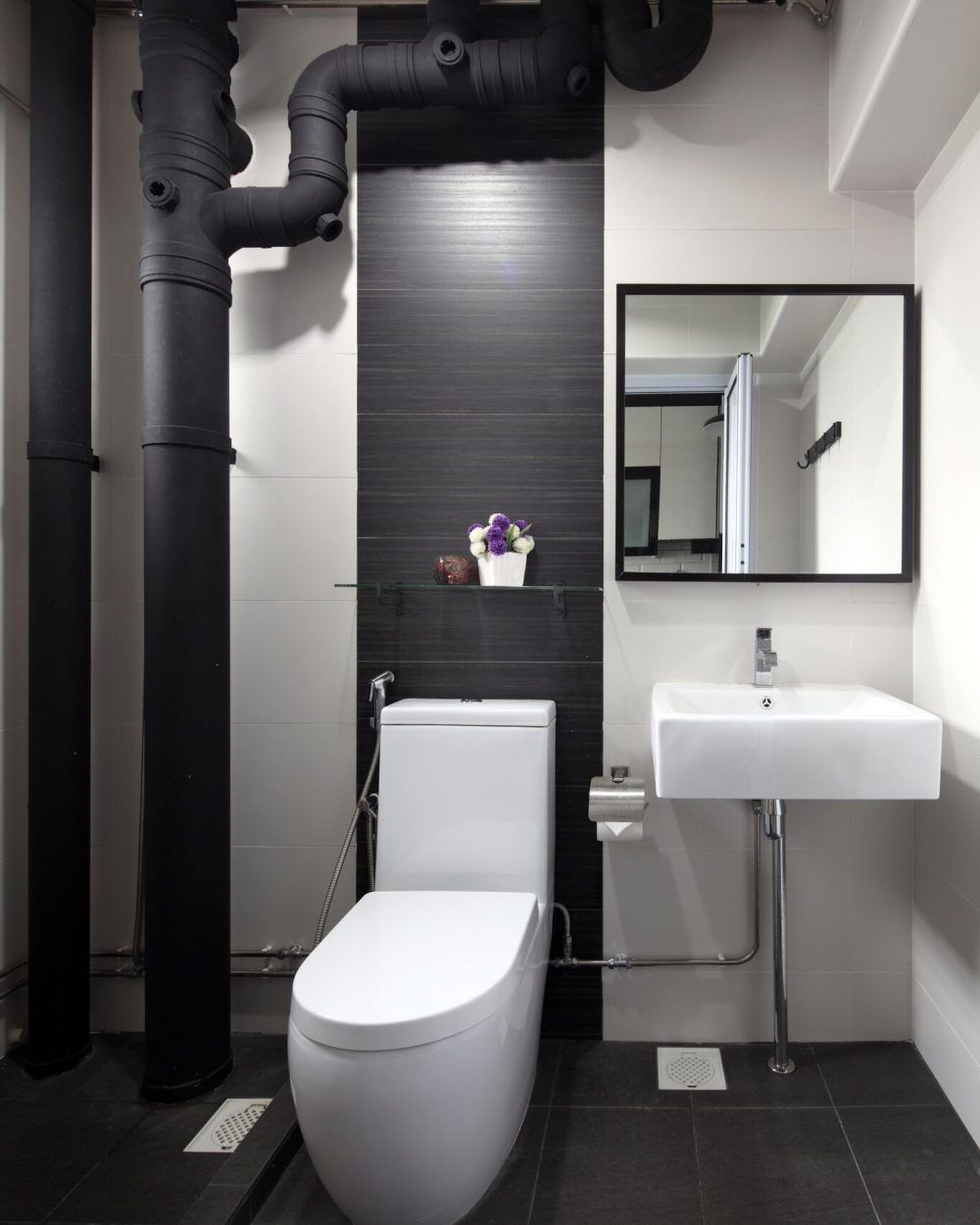 Exposed black pipework is left visible as a design feature against a dark feature tile wall, balanced by a crisp white sink and toilet.