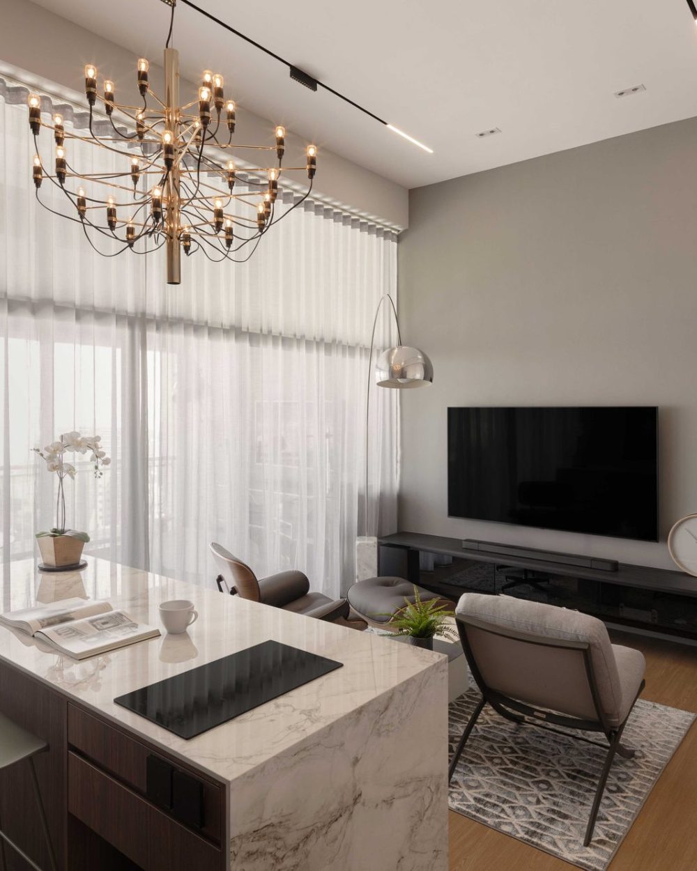 A tiered brass chandelier hangs over a marble-topped kitchen island doubling as a dining table, framed by sheer floor-to-ceiling curtains and a grey T