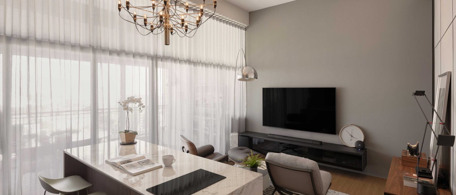 A tiered brass chandelier hangs over a marble-topped kitchen island doubling as a dining table, framed by sheer floor-to-ceiling curtains and a grey T