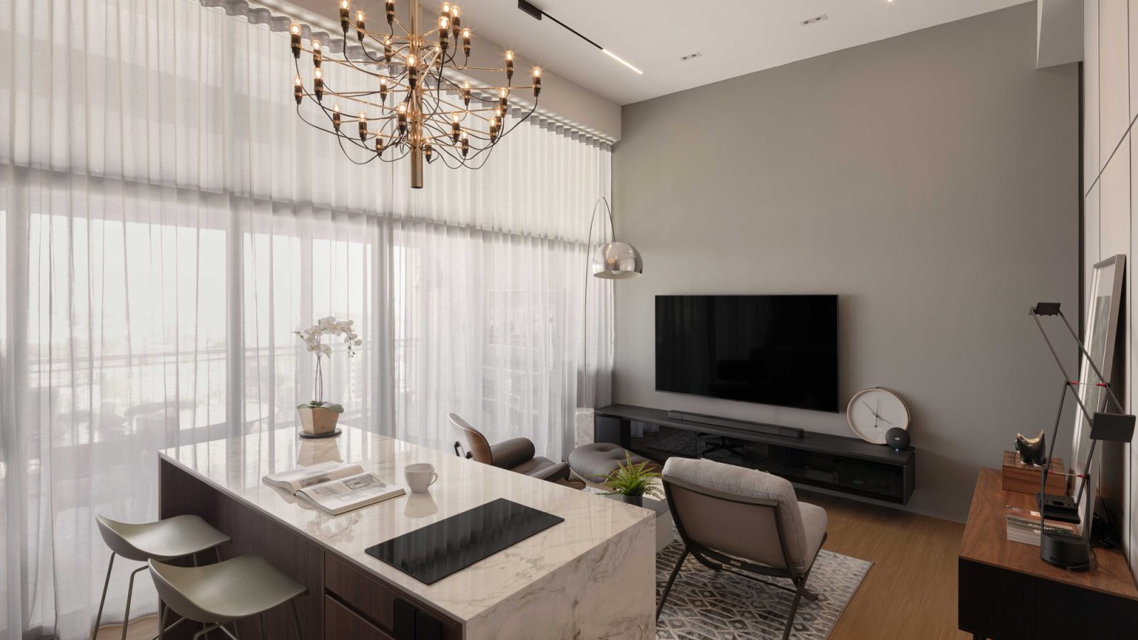 A tiered brass chandelier hangs over a marble-topped kitchen island doubling as a dining table, framed by sheer floor-to-ceiling curtains and a grey T