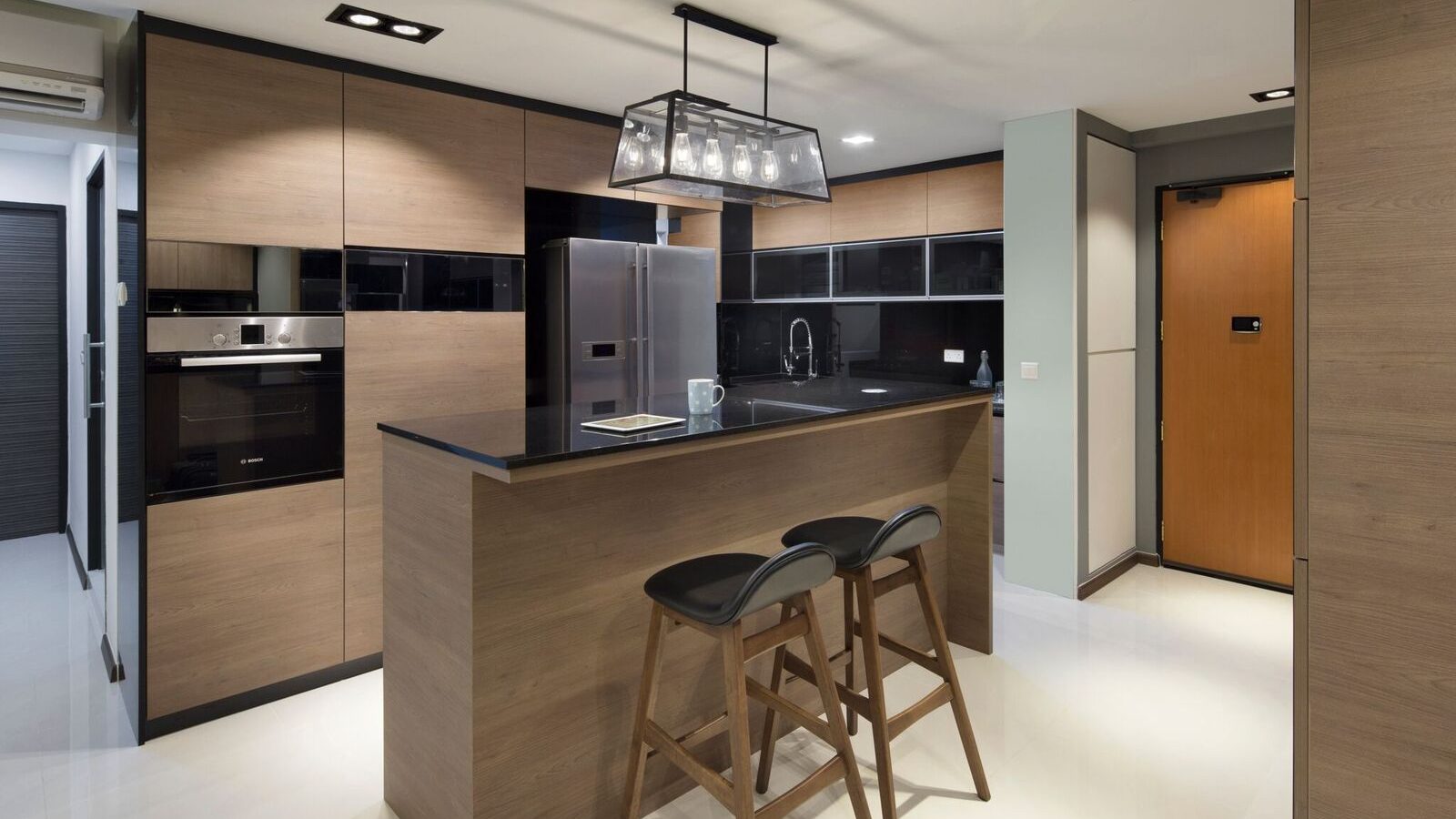 A boxy black pendant hangs over the kitchen island, framed by warm timber cabinetry and a glossy black upper unit run leading to the front door.