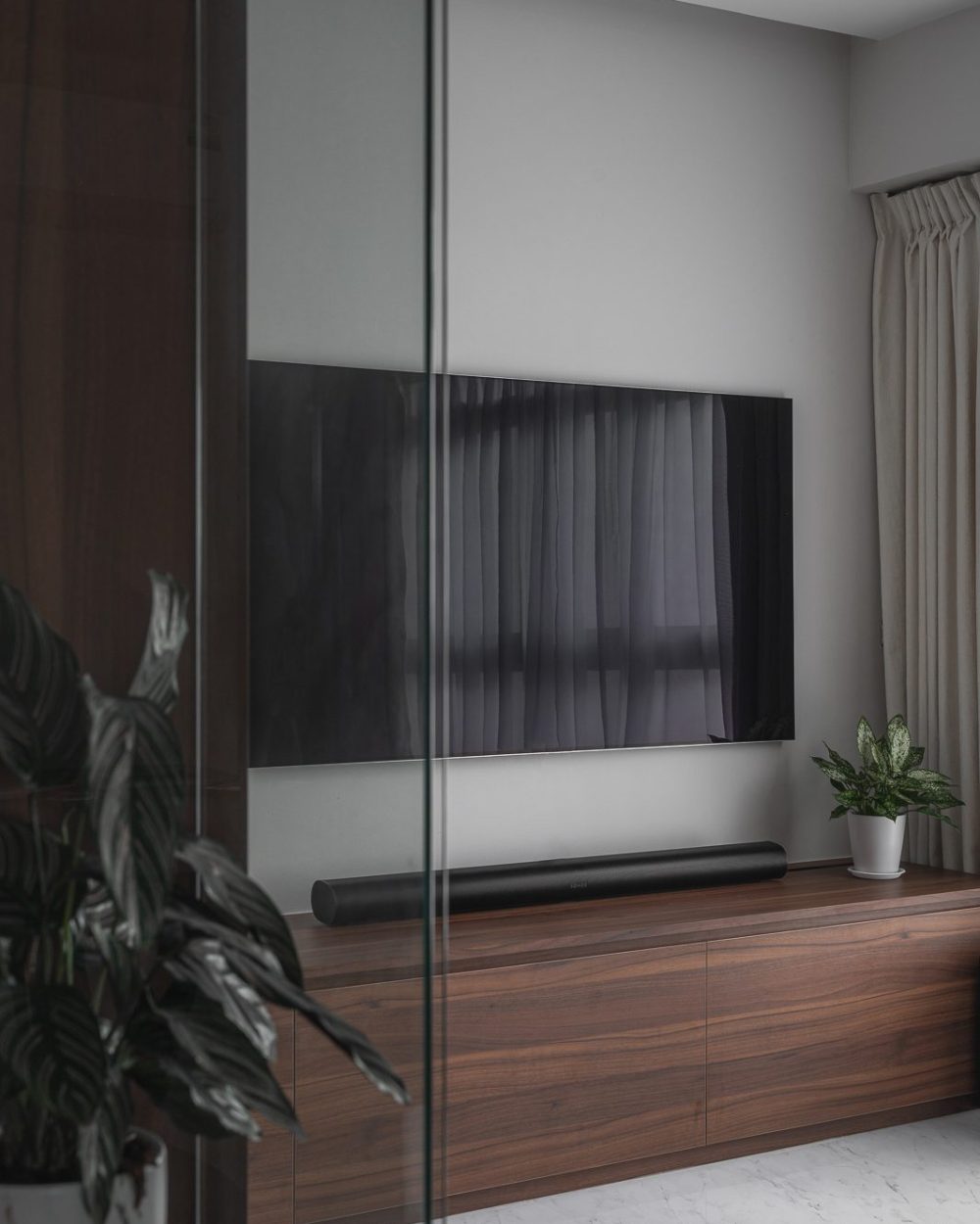 The glass door's reflection adds depth to the living room, layering the TV wall against the sheer curtained window.