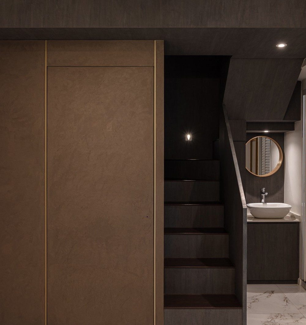 A textured timber sliding door sits beside a dark staircase leading up to a loft level, with a vanity basin and round mirror tucked beneath the stairs
