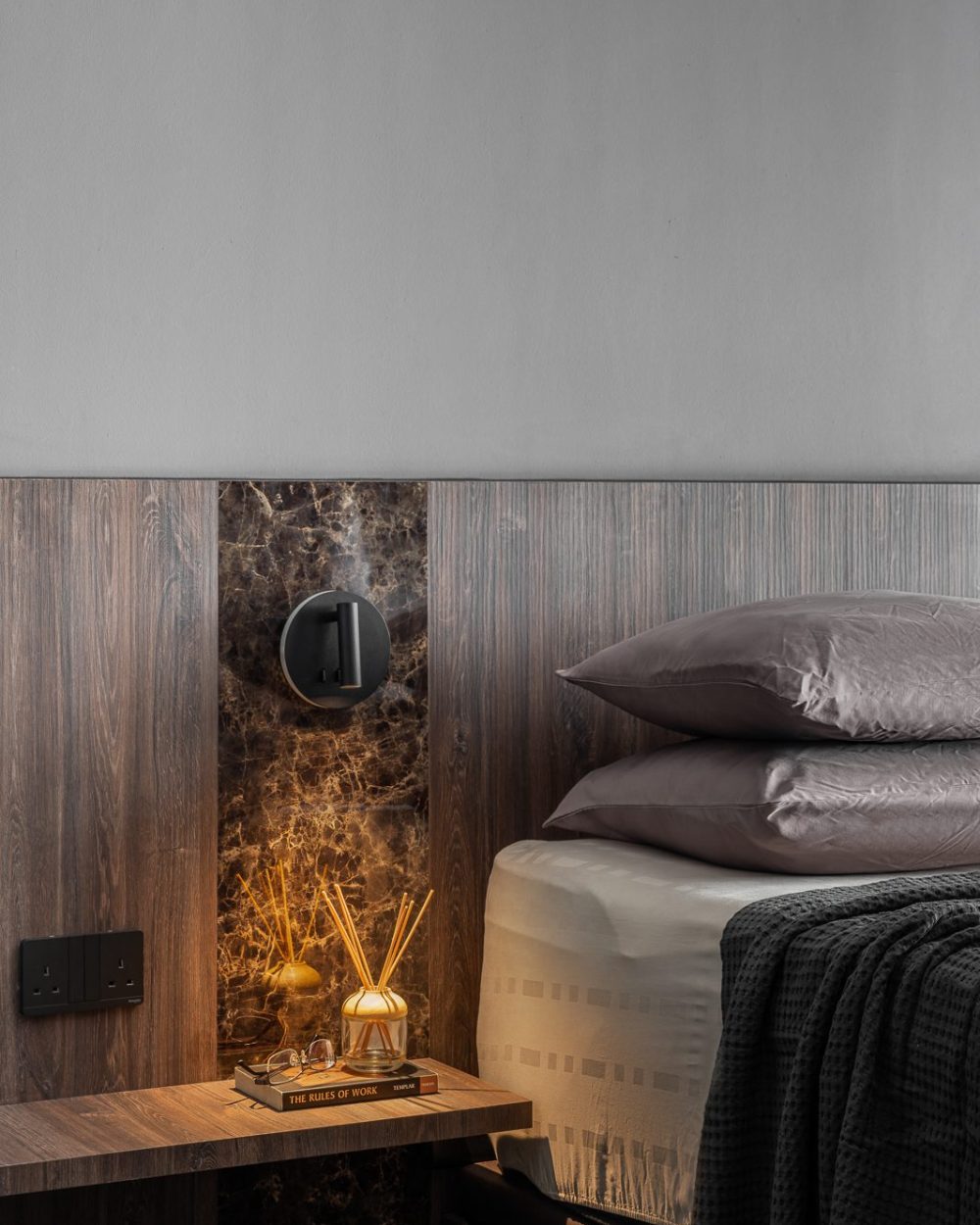 A dark marble panel and a sculptural black sconce sit beside the bed, styled with a reed diffuser and a favourite book.
