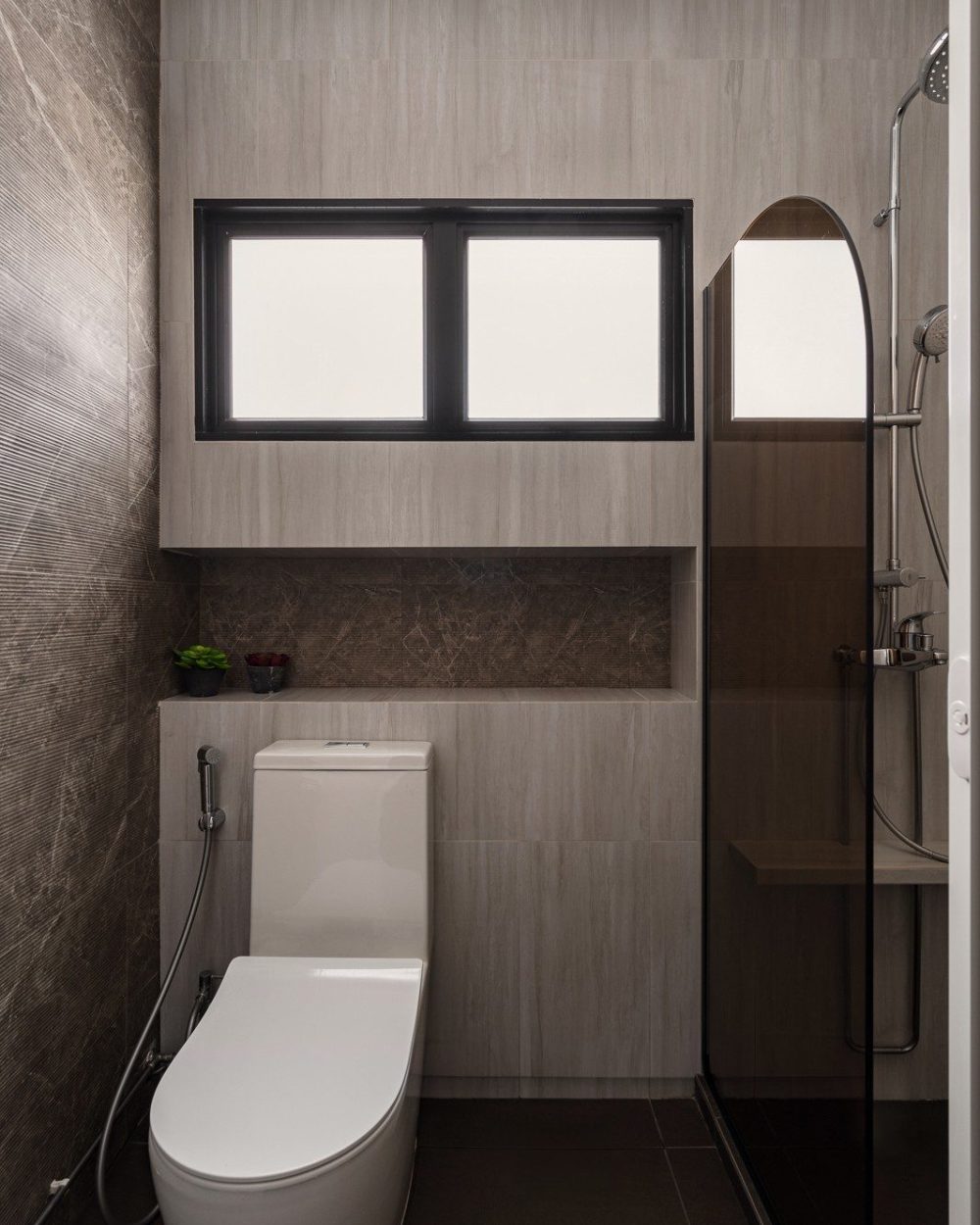 A window brings natural light into the ensuite, fitted with a wall-hung toilet, a tiled niche for toiletries, and a glass-screened shower with an arch