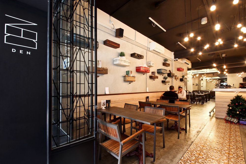 A simple line-drawn 'DEN' logo marks the black entrance wall, paired with a chalkboard easel listing the day's specials.