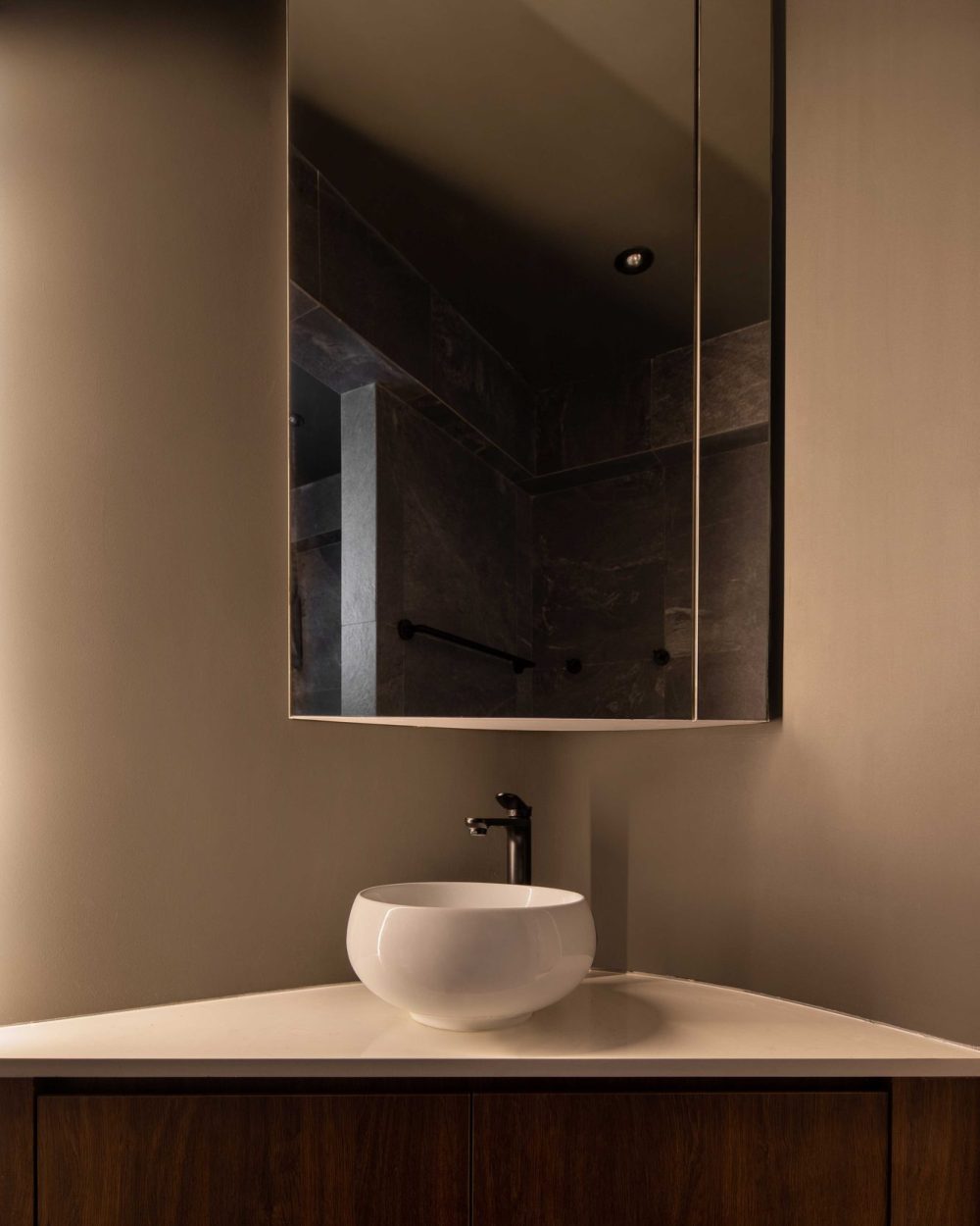 Dark stone tiling wraps the shower, reflected in a mirrored cabinet above a round vessel basin and matte black tap.