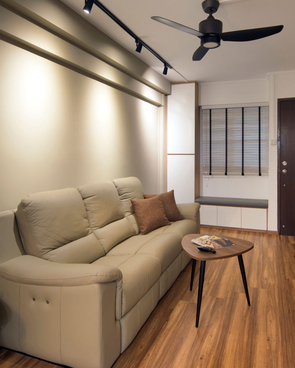 A beige leather sofa and round side tables sit against a soft grey feature wall, with a built-in storage bench tucked beneath the window.